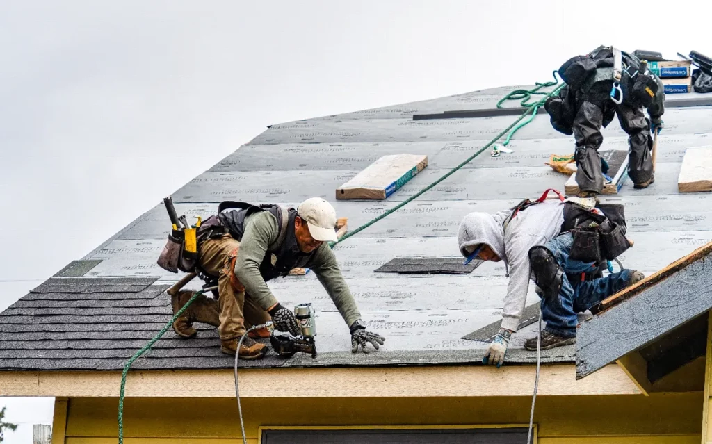 Zdjęcie przedstawia trzech pracowników montujących dachówki na dachu. Są wyposażeni w uprzęże bezpieczeństwa i różne narzędzia. Pracownicy są w trakcie układania dachówek, z jednym pracownikiem używającym gwoździarki do przymocowania dachówek na miejscu.