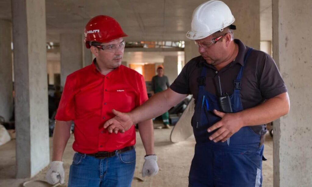 Na zdjęciu widać dwóch mężczyzn prowadzących rozmowę na placu budowy. Mężczyzna po lewej stronie ma na sobie czerwony kask i czerwoną koszulkę, a także niebieskie jeansy i białe rękawice oraz okulary ochronne. Mężczyzna po prawej nosi biały kask, ciemną koszulę oraz niebieskie ogrodniczki, do których z przodu przymocowano krótkofalówkę. W tle można dostrzec betonowe filary oraz jeszcze jednego pracownika w oddali.