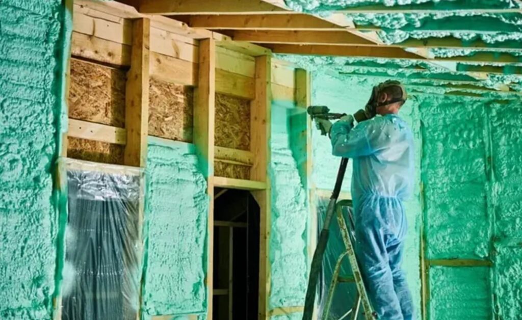 Na zdjęciu widzimy osobę ubraną w ochronny kombinezon i maskę, stojącą na drabinie. Osoba ta rozpyla zieloną piankę izolacyjną na ściany oraz sufit drewnianej konstrukcji.