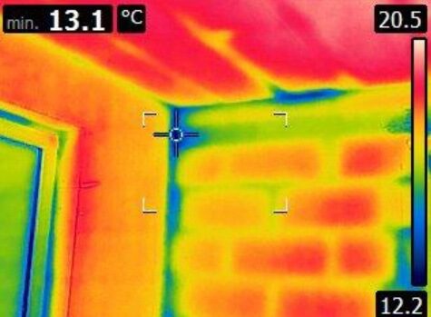 Obraz przedstawia widok z kamery termowizyjnej ukazujący róg pokoju. Kolory na termogramie odzwierciedlają różnice temperatur – czerwony i żółty oznaczają cieplejsze obszary, a niebieski i zielony wskazują chłodniejsze miejsca.