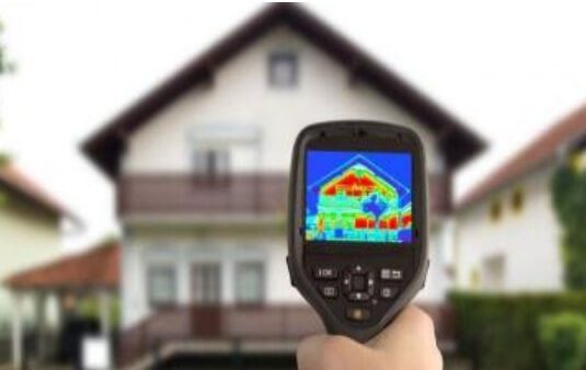 Na zdjęciu widzimy rękę trzymającą kamerę termowizyjną skierowaną na dom. Na ekranie kamery widoczna jest mapa cieplna budynku, wskazująca obszary o różnych temperaturach. Kolory na mapie pokazują różnice w rozkładzie ciepła - czerwone i żółte obszary prawdopodobnie reprezentują miejsca o wyższej temperaturze, a niebieskie i zielone wskazują chłodniejsze rejony.