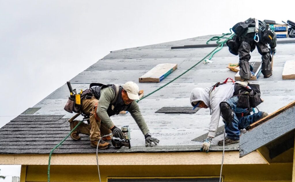 Na zdjęciu widać trzech pracowników montujących materiały dachowe na budynku. Są wyposażeni w uprzęże bezpieczeństwa oraz narzędzia, takie jak pistolety do gwoździ i pasy narzędziowe. Dach jest częściowo pokryty podkładem, a pracownicy przytwierdzają gont. 