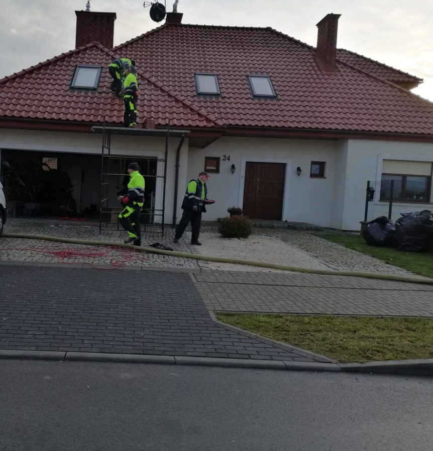 Na zdjęciu widzimy czterech pracowników ubranych w odblaskowe ubrania robocze, wykonujących prace na dachu domu pokrytego czerwonymi dachówkami. Jeden z nich stoi na rusztowaniu, drugi pracuje bezpośrednio na dachu, a trzeci i czwarty znajdują się na ziemi — każdy zaangażowany w inne zadanie.