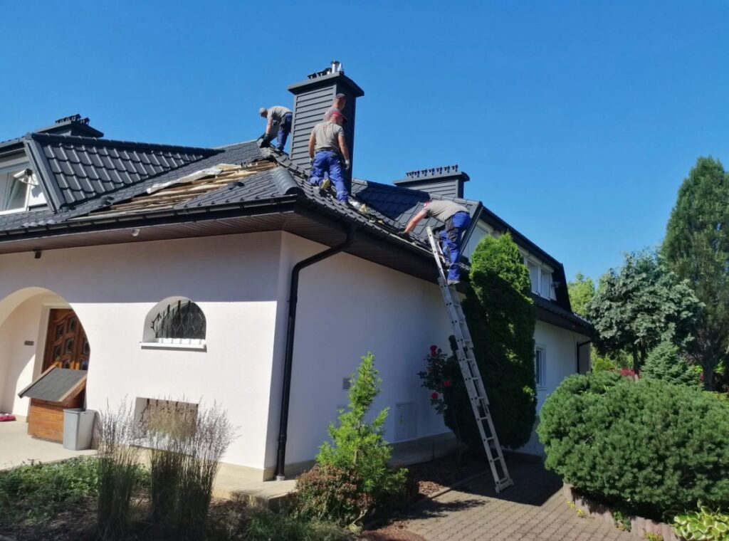 Na zdjęciu widzimy trzech pracowników wykonujących prace dekarskie na dachu domu. Jeden z nich stoi na drabinie, a dwóch pozostałych pracuje bezpośrednio na dachu, który wygląda na częściowo zdemontowany — widoczne są drewniane belki konstrukcyjne. Dom ma biały tynk, drewniane drzwi frontowe oraz kilka okien. Otoczenie jest schludne, z zielenią w postaci krzewów i roślin ozdobnych. Nad całą sceną rozciąga się bezchmurne, niebieskie niebo.
