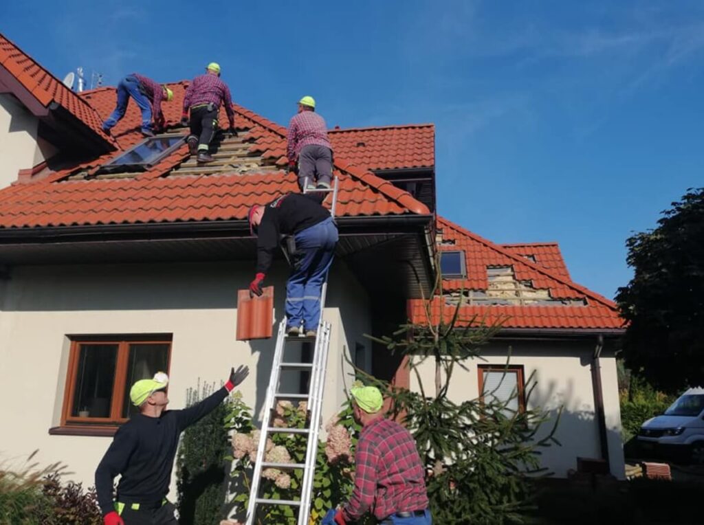 Zdjęcie przedstawia grupę mężczyzn w kaskach, którzy pracują na dachu domu jednorodzinnego. Dach jest pokryty czerwoną dachówką, ale widać uszkodzenia i brakujące elementy, co sugeruje, że trwa jego naprawa. Dwóch mężczyzn stoi na dachu, a jeden schodzi po drabinie, niosąc dachówkę. Czwarty mężczyzna, stojący na ziemi, wskazuje coś w górę, a piąty, ubrany w koszulę w kratę, stoi u podnóża drabiny. W tle widać fragment błękitnego, bezchmurnego nieba oraz fragment samochodu dostawczego po prawej stronie.