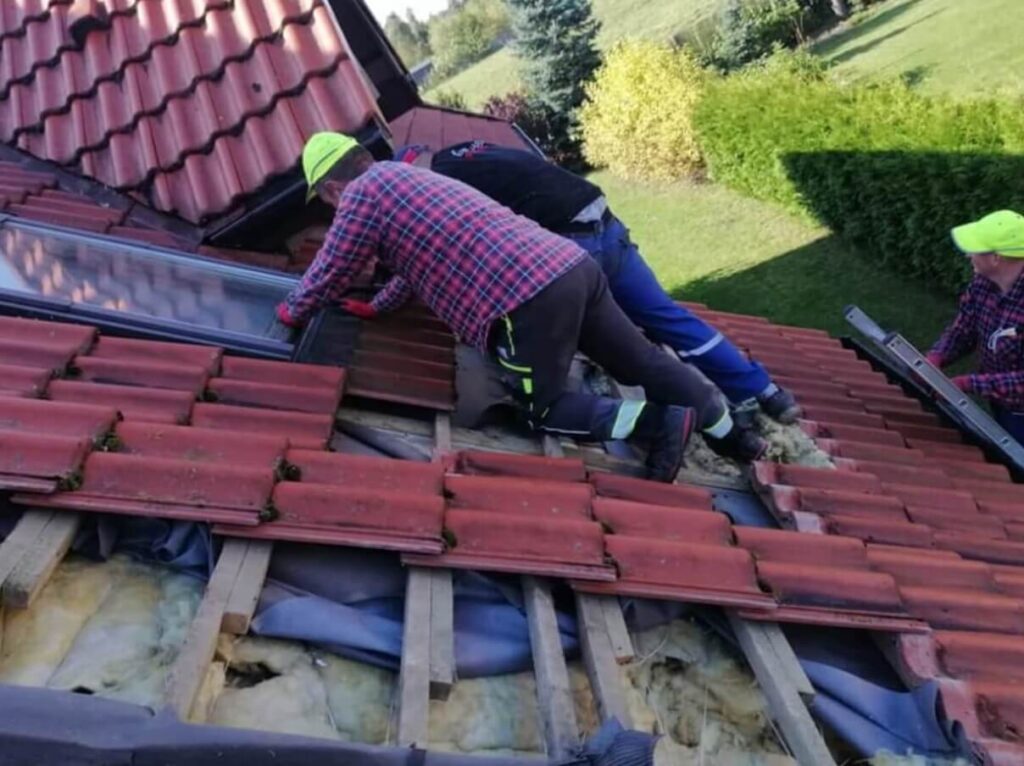 Na zdjęciu widzimy trzech pracowników budowlanych wykonujących prace na dachu domu pokrytego czerwoną dachówką. Część dachówek została usunięta, odsłaniając drewnianą konstrukcję i warstwę izolacyjną, co sugeruje trwający remont. Jeden z robotników klęczy przy świetliku, drugi mu asystuje, a trzeci trzyma drabinę, dbając o bezpieczeństwo i dostęp. Wszyscy mają na sobie odzież roboczą i jaskrawe czapki ochronne, co podkreśla przestrzeganie zasad BHP. W tle widać zielone trawniki, drzewa.