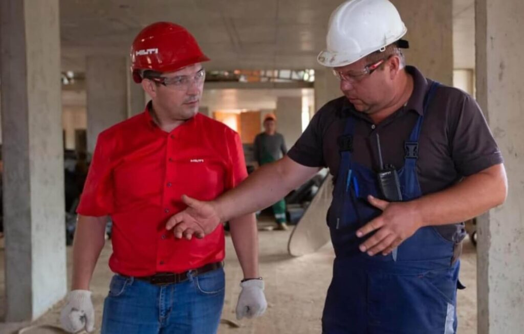 Na zdjęciu widzimy dwóch pracowników budowlanych prowadzących rozmowę na terenie aktywnej budowy. Jeden z nich, ubrany w czerwony kask i koszulkę z logo „HILTI”, słucha uważnie, podczas gdy drugi — w białym kasku, ciemnej koszuli i niebieskich ogrodniczkach — gestykuluje, prawdopodobnie wyjaśniając szczegóły techniczne. W tle widać betonowe filary, materiały budowlane oraz trzeciego pracownika w pomarańczowym kasku.