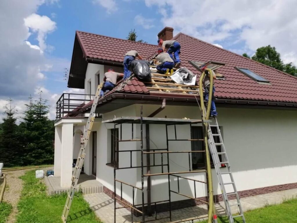 Na zdjęciu widzimy grupę pracowników budowlanych zajętych naprawą dachu dwupiętrowego domu o białych ścianach i czerwonej dachówce. Robotnicy pracują na różnych poziomach — część z nich znajduje się bezpośrednio na dachu, inni korzystają z rusztowania i drabin. W użyciu są drewniane belki i czarna folia, co sugeruje etap zabezpieczania konstrukcji przed warunkami atmosferycznymi. Tło tworzy naturalne otoczenie z drzewami i częściowo zachmurzonym niebem.