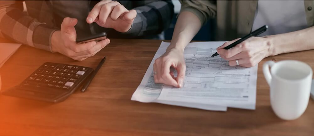 Na zdjęciu widzimy dwie osoby siedzące przy drewnianym stole, skupione na pracy z dokumentami finansowymi. Jedna z nich trzyma długopis i wskazuje na wydrukowany formularz -zestawienie budżetowe. Druga osoba gestykuluje, trzymając w dłoni smartfon, jakby odnosiła się do danych lub wyjaśniała coś na podstawie informacji z ekranu.