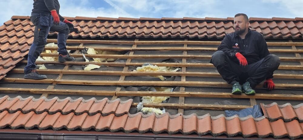 Na zdjęciu widzimy dwóch pracowników  w ciemnych ubraniach, zajmujących się remontem dachu. Jeden z nich stoi na odsłoniętych drewnianych łatach, drugi siedzi. Dach jest częściowo rozebrany — widoczne są warstwy izolacji i konstrukcja nośna.