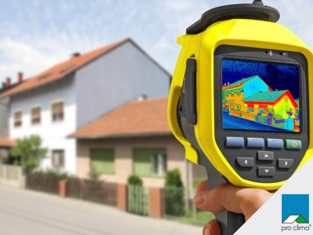 Na zdjęciu widzimy osobę trzymającą kamerę termowizyjną skierowaną na rząd domów. Na ekranie urządzenia wyświetla się obraz w kolorach odpowiadających różnicom temperatur: czerwienie, pomarańcze i żółcie wskazują cieplejsze obszary, a błękity i zielenie — chłodniejsze. Tło pokazuje te same budynki w świetle dziennym, co podkreśla kontrast między widzialnym a termicznym obrazem.