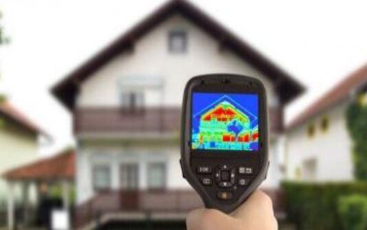 Na zdjęciu widzimy osobę trzymającą kamerę termowizyjną skierowaną na fasadę domu. Na ekranie urządzenia wyświetla się obraz termiczny, ukazujący rozkład temperatur na powierzchni budynku — cieplejsze obszary zaznaczone są czerwienią i żółcią, chłodniejsze błękitem i zielenią.