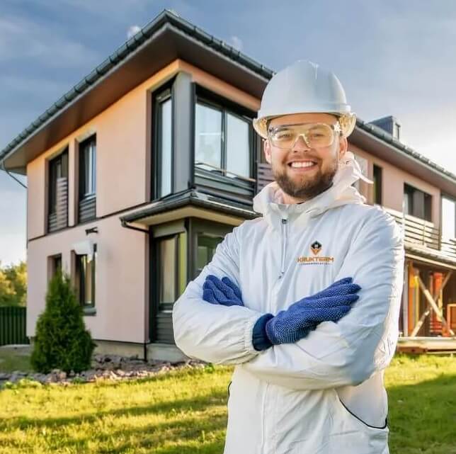 Na zdjęciu widać uśmiechniętego mężczyznę stojącego przed nowoczesnym domem jednorodzinnym. Ma na sobie biały kombinezon roboczy z logo firmy, kask ochronny oraz przezroczyste okulary zabezpieczające. Jego ręce są skrzyżowane na klatce piersiowej, a na dłoniach ma niebieskie rękawice robocze. W tle znajduje się dwukondygnacyjny budynek o nowoczesnej bryle, jasnej elewacji i dużych przeszkleniach.