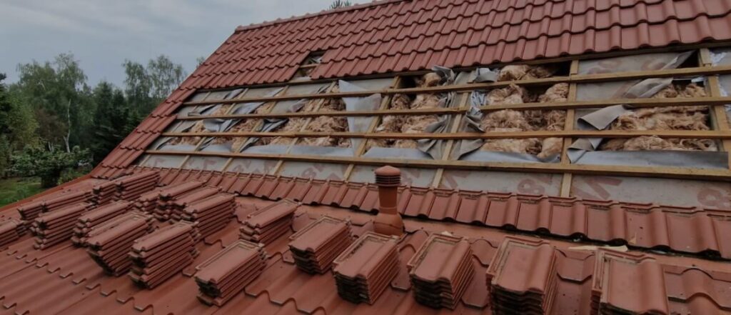 Na zdjęciu widać dach budynku w trakcie prac remontowych lub modernizacji izolacji. Część dachówek została zdjęta, odsłaniając warstwy konstrukcyjne oraz materiał ociepleniowy znajdujący się pod pokryciem. Na pierwszym planie ułożone są stosy dachówek przygotowanych do ponownego montażu.